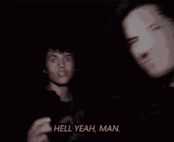 a man is talking into a microphone in a dark room while another man is smoking a cigarette in the background .