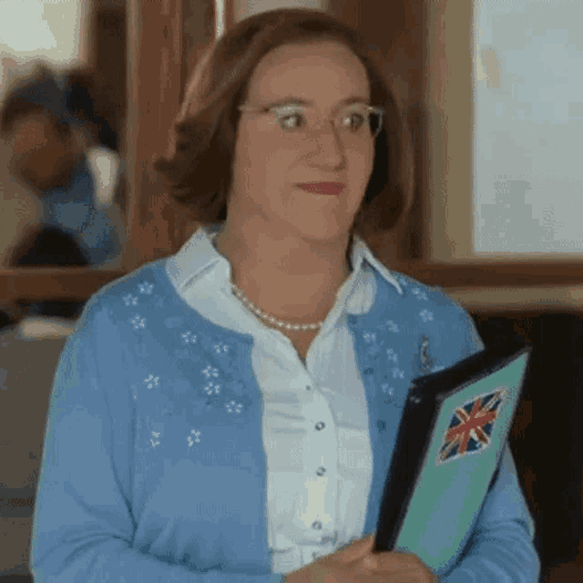 a woman wearing glasses and a blue cardigan is holding a folder with a british flag on it .