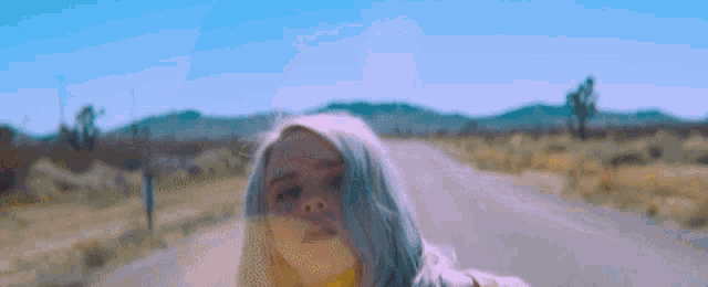 billie eilish is walking down a desert road wearing a yellow shirt .
