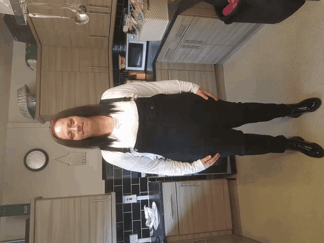 a woman in overalls stands in a kitchen with a clock on the wall that says 10:08