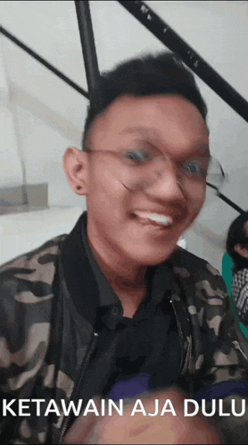 a man wearing glasses and a camouflage jacket smiles for the camera with the words ketawain aja dulu below him