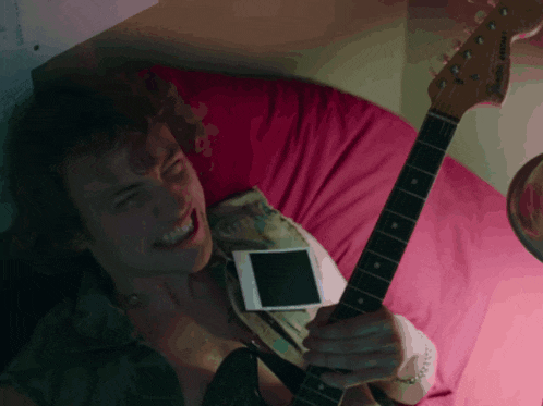 a man laying on a bed with a guitar and a cell phone