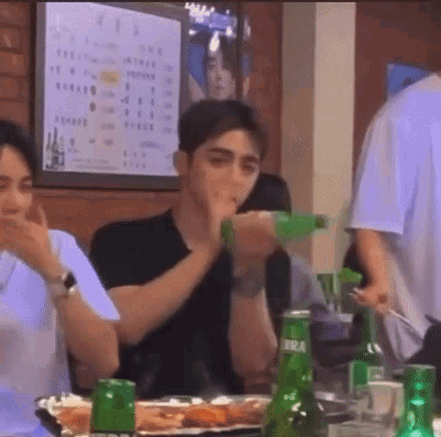 a man is drinking from a green bottle while sitting at a table in a restaurant .