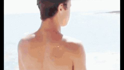 the back of a shirtless man standing on the beach looking at the ocean .