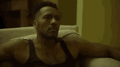 a man in a black tank top is sitting on a couch looking at the camera .