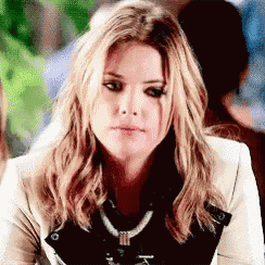 a woman with long blonde hair and a necklace is sitting at a table with a serious look on her face .