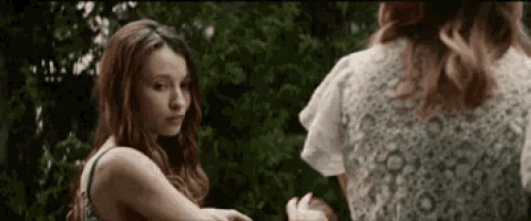 two women are standing next to each other in front of trees . one of the women is wearing a white lace top .
