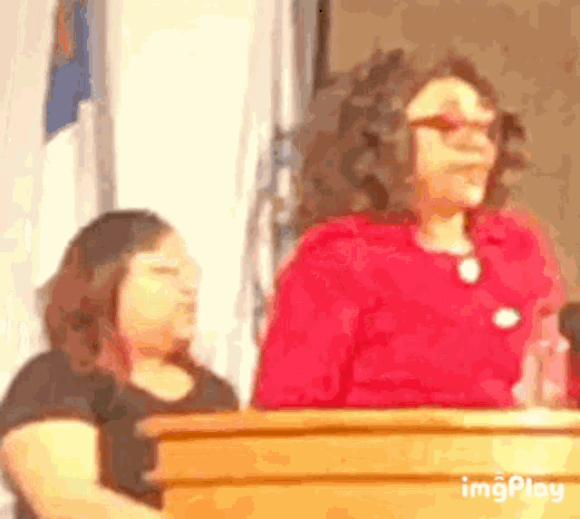 a woman is standing at a podium giving a speech while another woman sits behind her .
