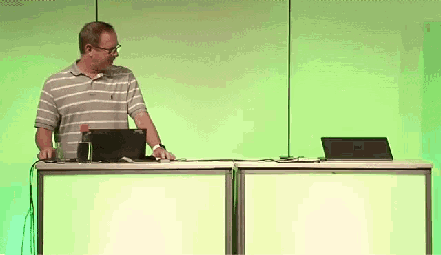 a man in a striped shirt stands at a desk with a laptop and a bottle of water