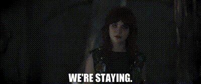 a woman in a black dress is standing in a dark room and says `` we 're staying . ''