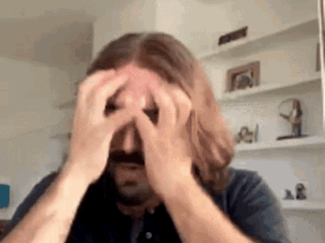 a man with long hair and a beard is rubbing his forehead with his hands .