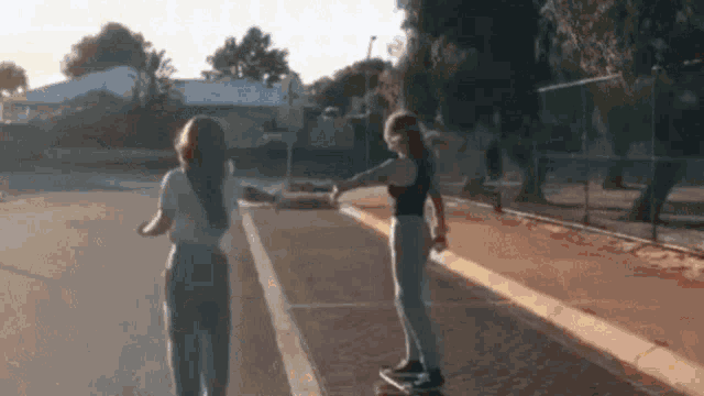 two girls are standing next to each other on a sidewalk while one is riding a skateboard .