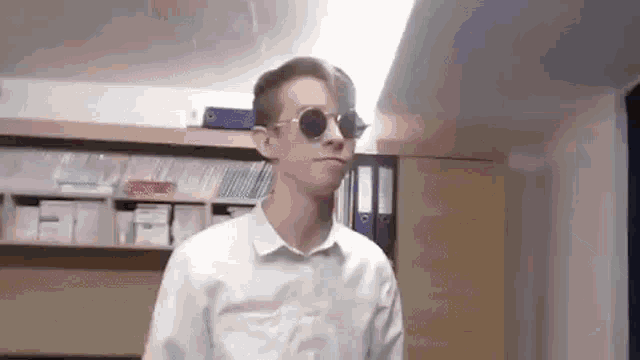 a man wearing sunglasses and a white shirt is standing in front of a bookshelf .