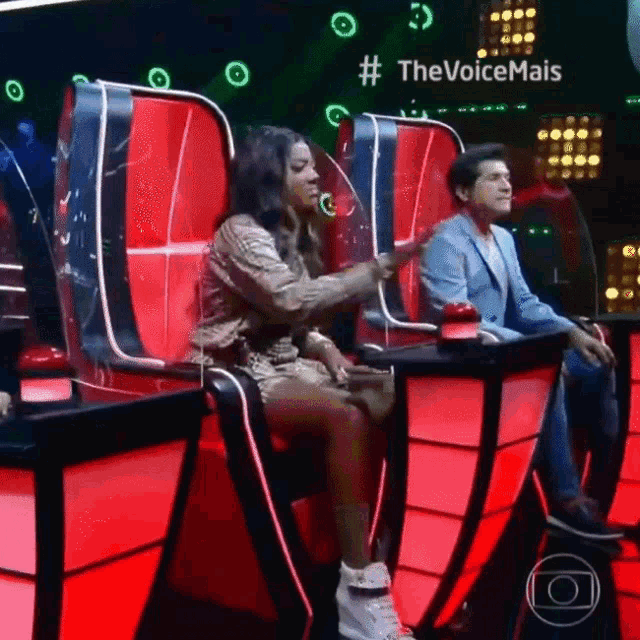 a woman is sitting in a chair with a man sitting behind her and a sign that says # thevoicemais