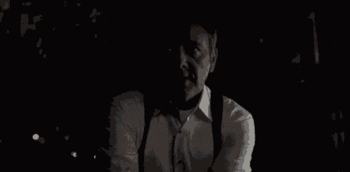 a man in a white shirt and suspenders is sitting in the dark and saying no more pain .