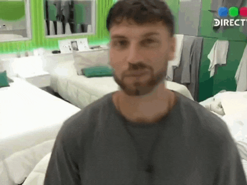 a man with a beard is standing in front of a bed in a room with a direct sign on the wall