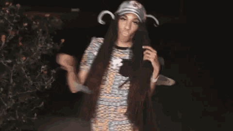 a woman with long hair wearing a hat and a t-shirt is standing in front of a bush .