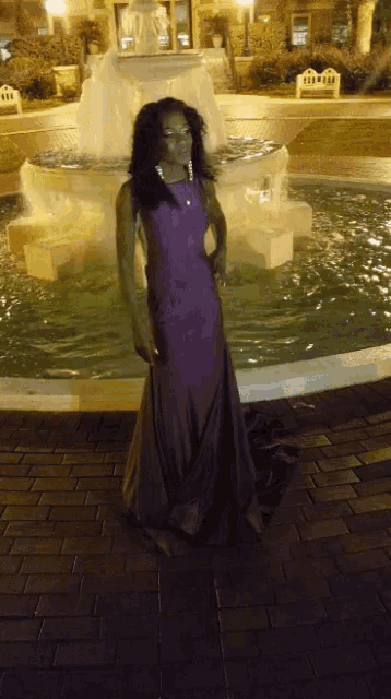 a woman in a long purple dress is standing in front of a fountain