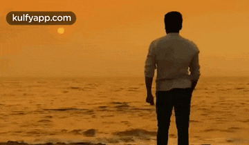 a man is standing on a beach looking out over the ocean at sunset .