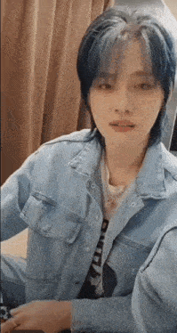 a young man with blue hair is wearing a denim jacket and sitting at a table .