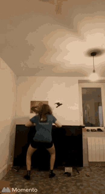 a woman in a blue shirt is squatting in a room with a momento logo on the bottom right