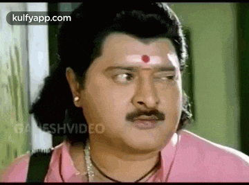 a man with a mustache and a red dot on his forehead is wearing a pink shirt and necklace .