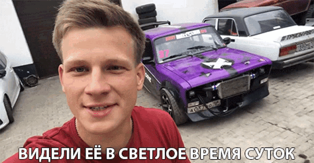 a man in a red shirt is standing in front of a purple car with the number 37 on the windshield