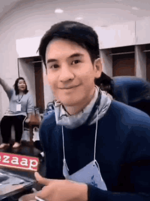 a man wearing a blue sweater and a mask is smiling while a woman sits on a chair in the background .