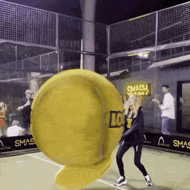 a woman stands on a tennis court with a large yellow tennis ball that says smash on it