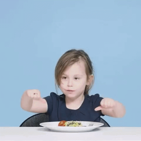 a little girl is sitting at a table with a plate of food and making a thumbs up sign .