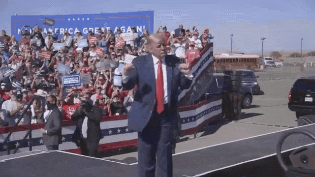 a man in a suit and tie stands on a stage in front of a sign that says america great again