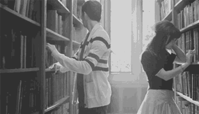 a man and a woman are standing next to each other in a library looking at books .