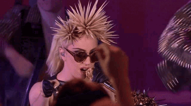 a woman wearing sunglasses and a spiked mohawk sings into a microphone
