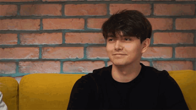 a young man sitting on a yellow couch with a brick wall behind him