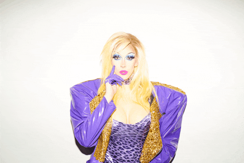 a woman wearing a purple jacket and leopard print dress holds her finger to her mouth