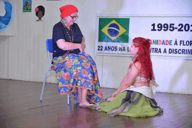 a woman sits on a chair next to a woman kneeling down in front of a sign that says 1995-20
