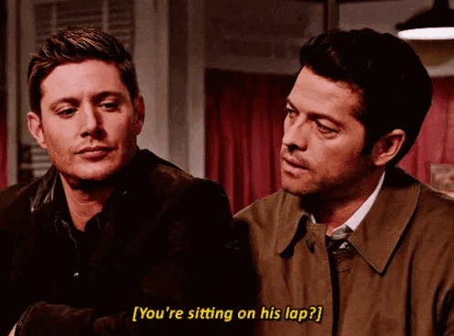 two men are sitting next to each other in a room and one of them is asking the other if he is sitting on his lap .