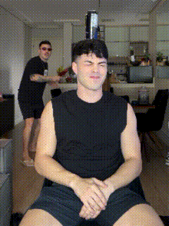 a man in a black tank top and shorts is sitting in a chair with his hands folded while another man stands behind him