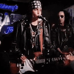 a man is singing into a microphone while another man plays a guitar in front of a johnny 's sign .
