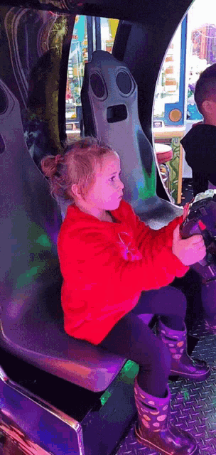 a little girl in a red sweatshirt is sitting in a chair holding a steering wheel