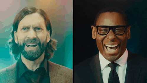 a man with a beard and glasses laughs next to another man in a suit