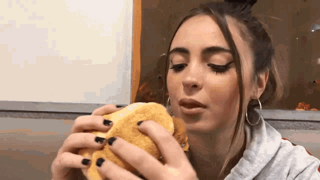 a woman with black nails is eating a hamburger in front of a window