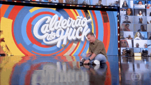 a man sits on the floor in front of a screen that says caldeirão do puck