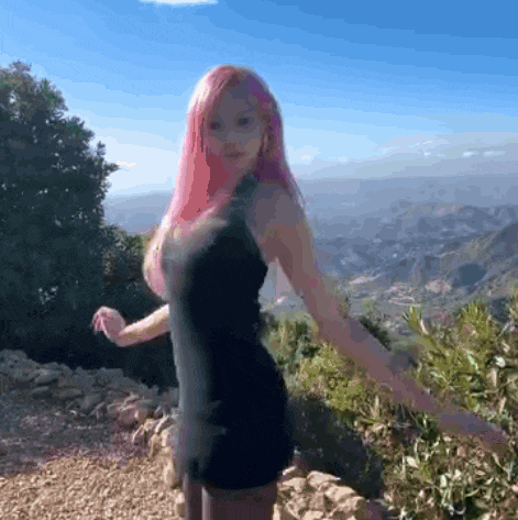 a woman with pink hair is wearing a black dress and standing on top of a hill .