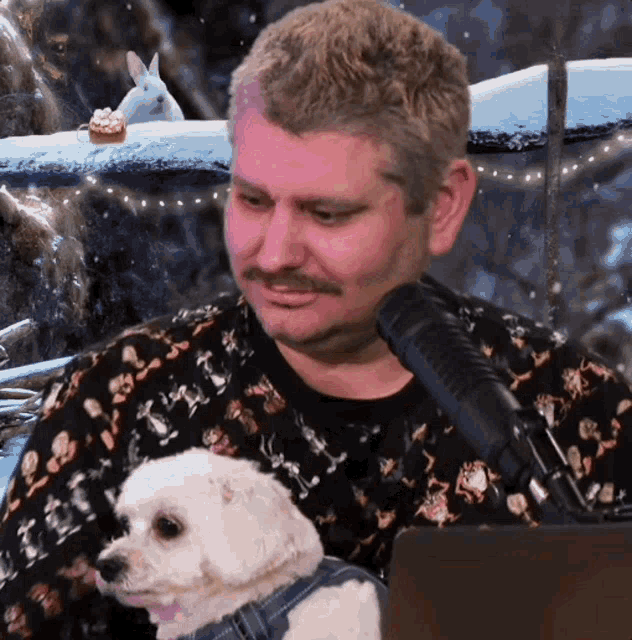 a man with a mustache holds a small white dog in front of a microphone