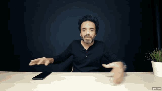 a man in a black shirt is sitting at a desk with his hands outstretched and a phone on the table