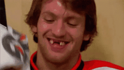 a man with missing teeth is smiling in a locker room while holding a hockey stick .