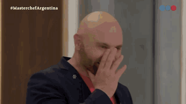 a bald man covering his face with his hand in front of a masterchef argentina sign