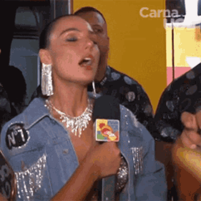 a woman in a denim jacket is talking into a microphone while wearing a necklace with the number 1 on it