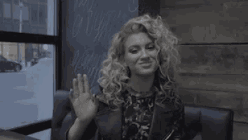 a woman with curly hair is sitting at a table in front of a window waving .
